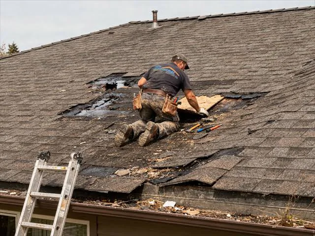 Roofer tearing damaged shingles off a roof section down to the bare decking during an emergency repair