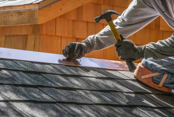Roofer hammering new metal flashing into place along a shingle roof against a shingled wall